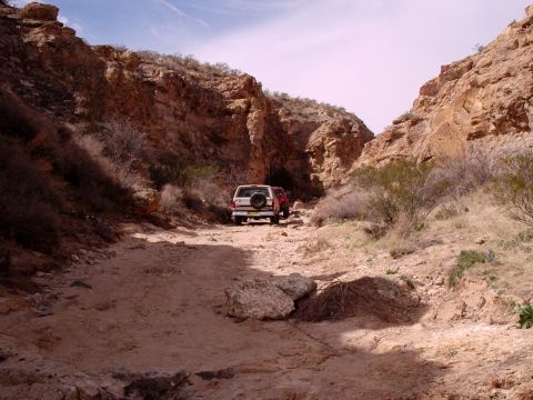 1996 Ford Bronco - Two SUVs, including a white 1996 Ford Bronco, navigate a narrow dirt trail through a rocky canyon.