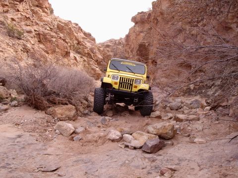 1996 Ford Bronco - Yellow Jeep Wrangler driving through a narrow, rocky canyon with sparse vegetation.