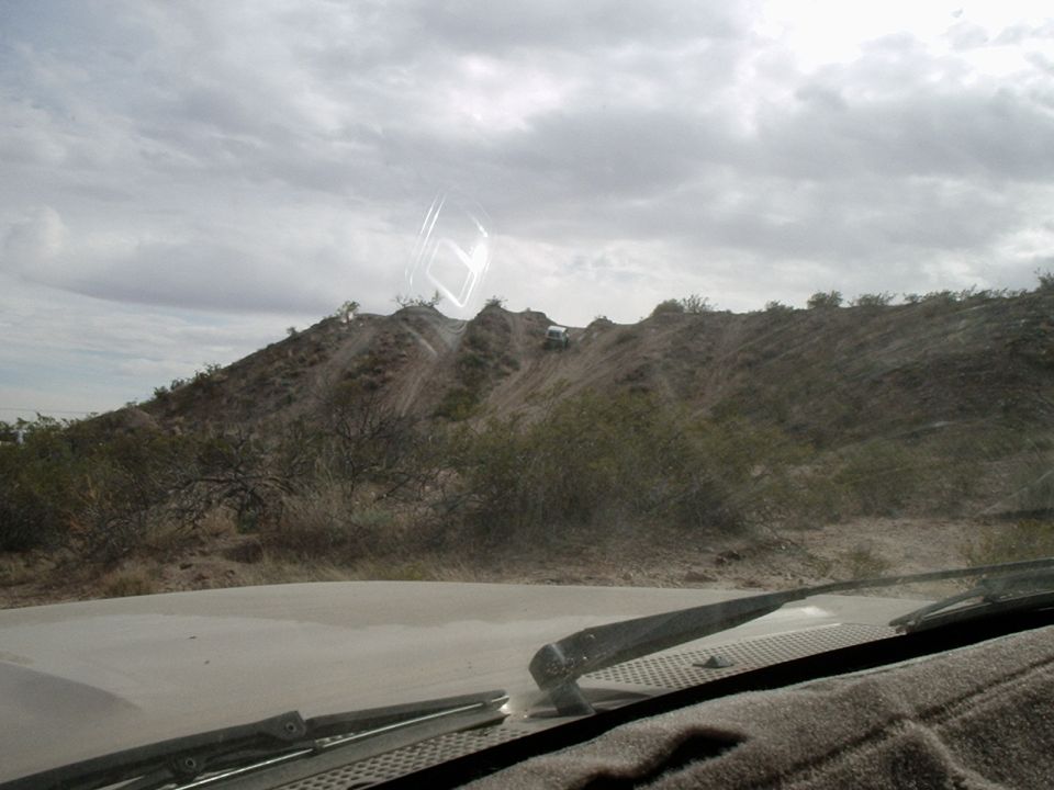 Two off-road vehicles ascend a steep, dusty hill under a cloudy sky. View from inside a vehicle