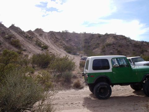 1996 Ford Bronco - Several off-road vehicles are parked on a sandy trail in a desert landscape with scrub brush and hills.