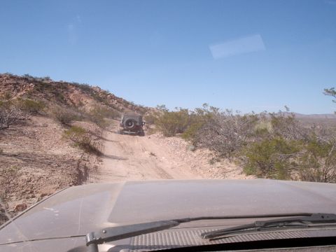 1996 Ford Bronco - Two vehicles navigate a rough dirt path through a dry, scrubby terrain under a clear blue sky.