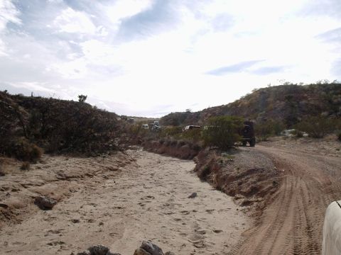 1996 Ford Bronco - Off-road vehicles are parked along a dirt road in a dry creek bed with hills in the background.