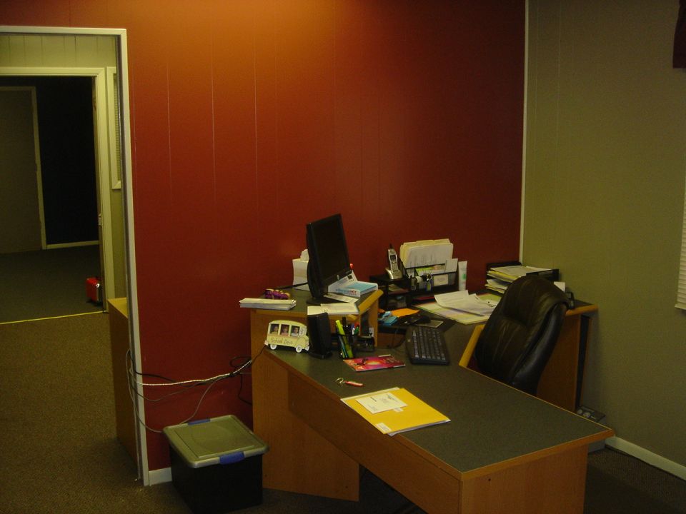 Office desk with monitor, keyboard, papers and black chair against a red wall