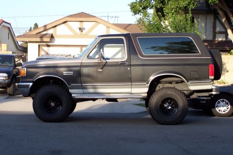 1990 Ford Bronco - A lifted Ford Bronco is parked on a residential street, showing its side profile with large off-road tires.