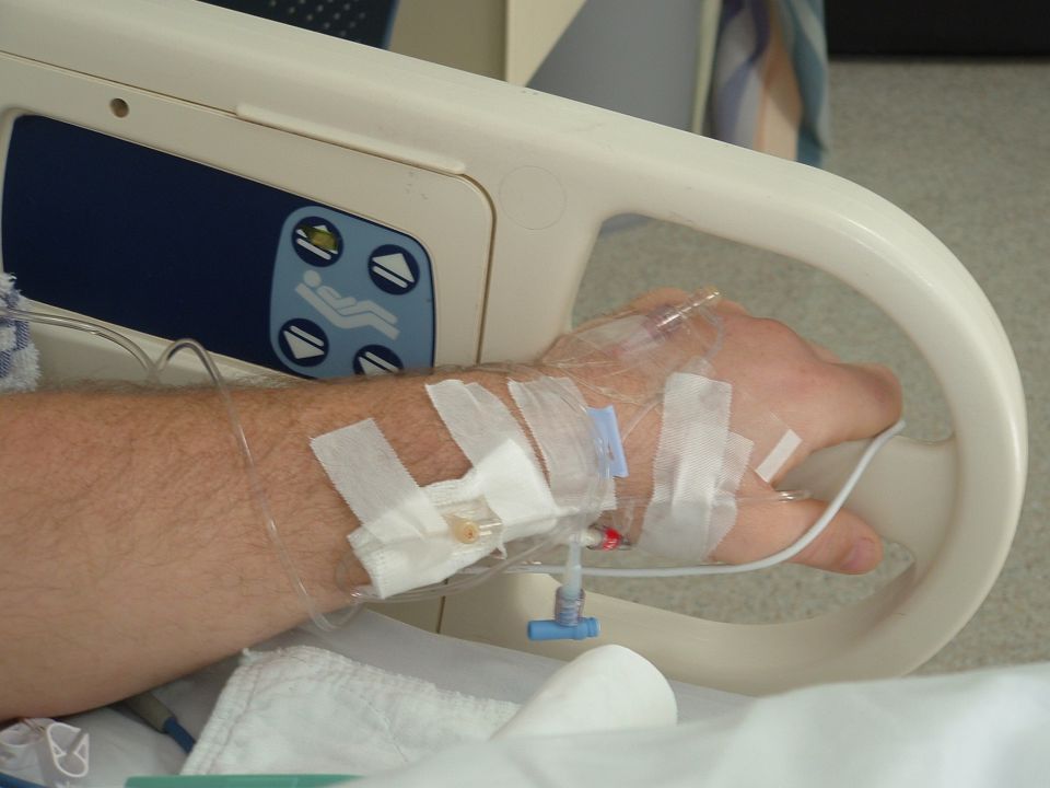 Arm on hospital bed rail with IV line taped to the hand; bed control buttons visible.