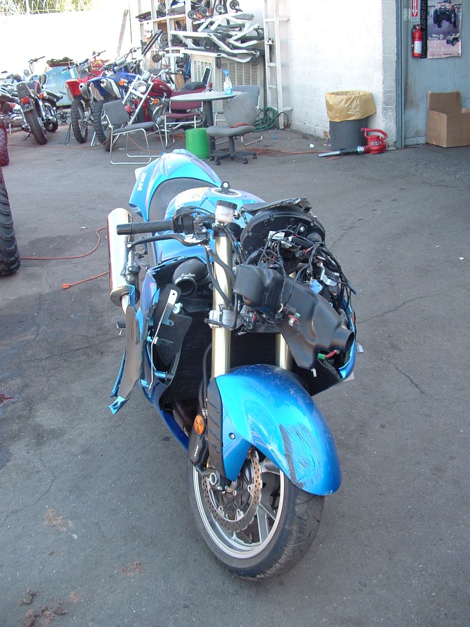 Front view of wrecked blue Kawasaki sport bike with fairing removed, exposed wiring and radiator, in a shop lot