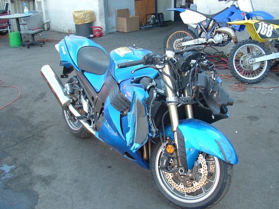 Blue Kawasaki ZX-14 with front fairing and headlight removed, showing crash damage, parked outside a shop