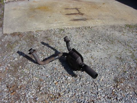 1991 Ford Bronco - A vehicle exhaust system part lies on a gravel surface next to a concrete pad.