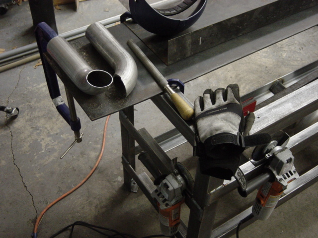 Metal exhaust pipe section clamped to a workbench with a file and work gloves