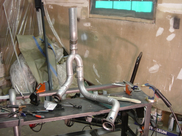 Tack-welded multi-bend exhaust assembly standing on a shop welding table with MIG torch and tools