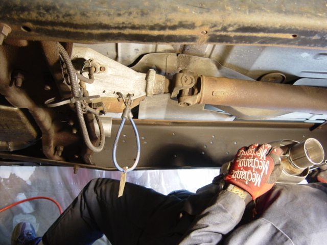 Person in Mechanix gloves test-fitting exhaust pipe under a truck near driveshaft and transfer case