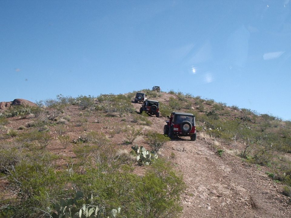 Several Jeeps driving up a rocky hill on a dirt path. Desert vegetation is visible