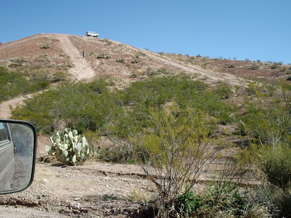White SUV parked on a dirt hill with a person standing nearby. Desert vegetation and a dirt road are visible