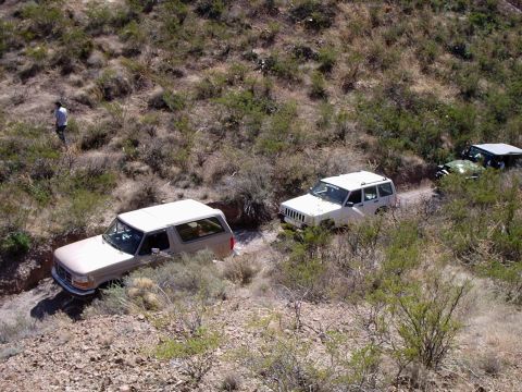 1996 Ford Bronco - A convoy of off-road vehicles, including a 1996 Ford Bronco and a Jeep Cherokee, traverses a dry, brush-lined trail.