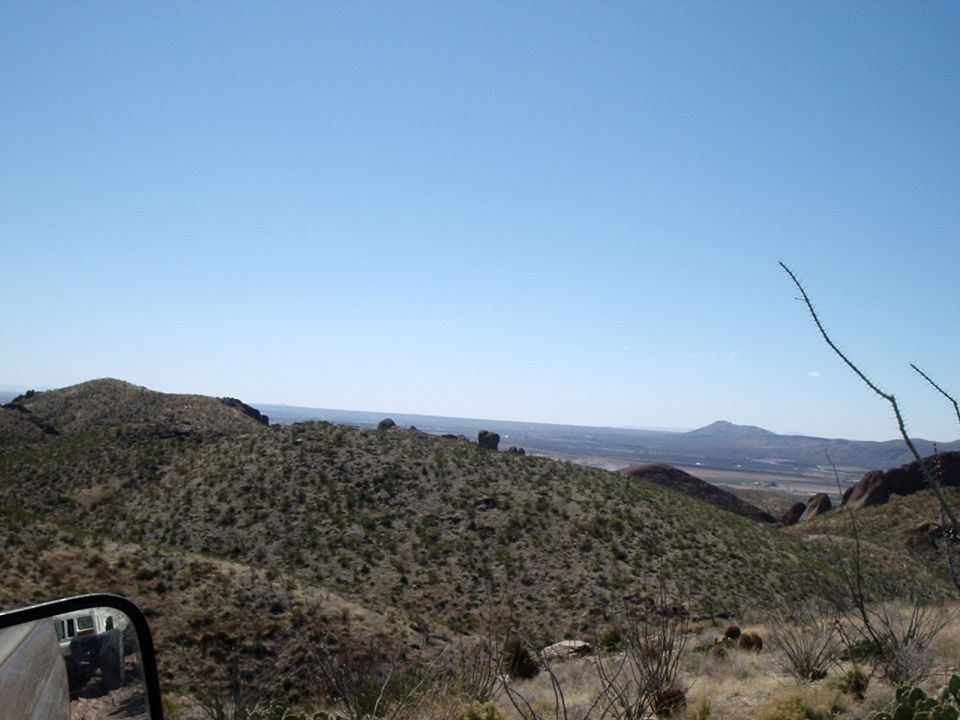 Desert landscape with scrub brush and mountains under a clear blue sky, side mirror reflection shows part of a vehicle