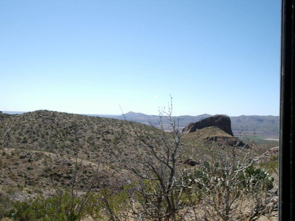 Desert landscape with hills, cacti, and a large rock formation under a clear blue sky