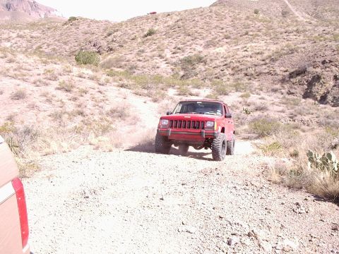 1996 Ford Bronco - A red Jeep Cherokee navigates a rough, off-road path in a desert landscape.