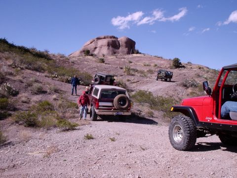 1996 Ford Bronco - Several off-road vehicles navigate a dusty trail with a large rock formation in the distance.