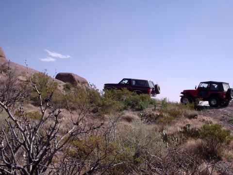 1996 Ford Bronco - Two off-road vehicles, a dark red SUV and a red Jeep, traverse a , arid landscape under a bright blue sky.