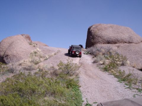 1996 Ford Bronco - A red Jeep Wrangler navigates a steep, gravel trail through a desert landscape with large rock formations.