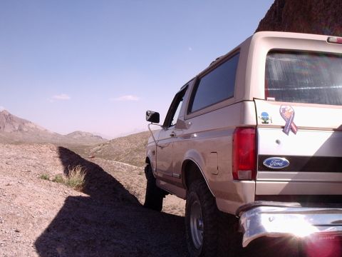 1996 Ford Bronco - Low-angle rear three-quarter view of a tan full-size Ford Bronco parked on a rocky desert hillside, with mountains and open scrub stretchin…
