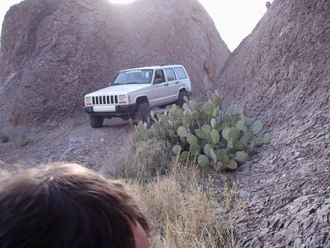 1996 Ford Bronco - A white Jeep Cherokee XJ navigates a , rocky terrain with sparse vegetation under a bright sky.