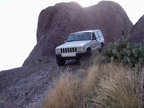 1996 Ford Bronco - A white Jeep Cherokee XJ navigates a rocky, off-road trail under a bright sky.