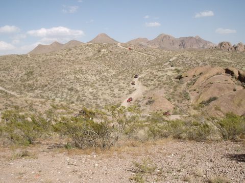 1996 Ford Bronco - Multiple off-road vehicles navigate a dusty trail in a mountainous desert terrain under a clear blue sky.