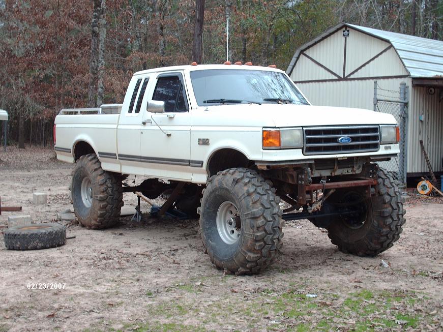White lifted Ford F-150 extended-cab pickup on huge mud tires parked by a barn
