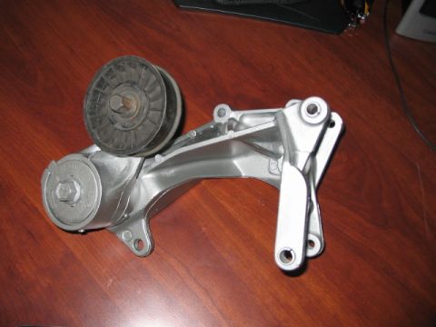 1991 Ford Bronco - A metal belt tensioner assembly with a pulley wheel and adjustment mechanism is shown on a wooden surface.