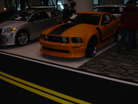 1996 Ford Crown Victoria - An orange Ford Mustang with black racing stripes is displayed at an auto show.