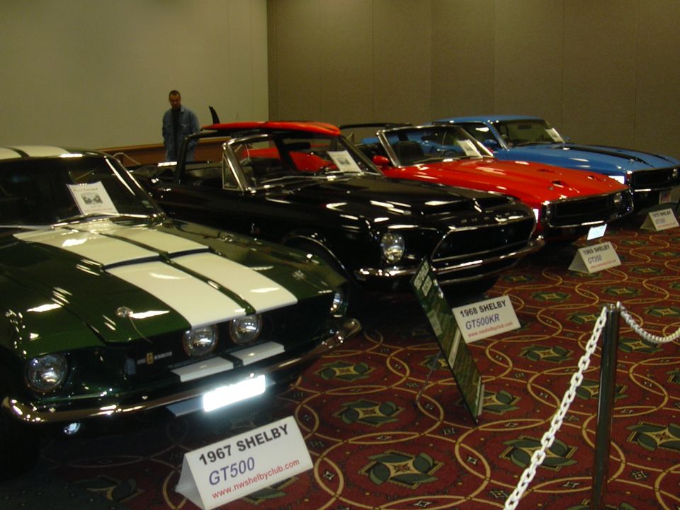 1967 Shelby GT500 with white stripes next to a black 1968 Shelby GT500KR, red and blue cars behind them