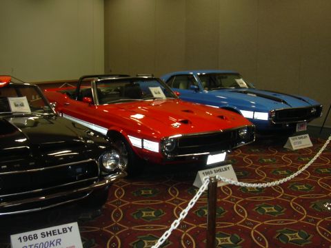1996 Ford Crown Victoria - Three classic Shelby Mustangs are displayed at an auto show, with a red convertible in the foreground.