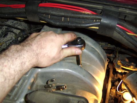 1989 Ford Bronco - A hand grips a socket wrench with an extension attached to a bolt on a gray engine component, working under the hood near bundled red and b…