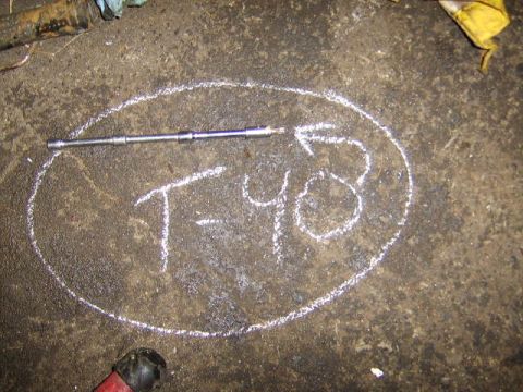 1989 Ford Bronco - A metal shaft or rod, possibly a torsion bar or shift linkage component, lies on a dirty concrete floor inside a chalk-drawn oval labeled '…