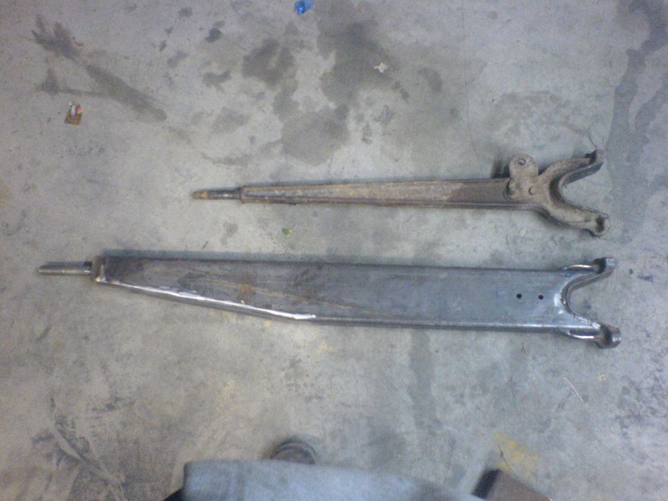 Two automotive suspension control arms, one rusty and one cleaned/painted, lie on a concrete floor