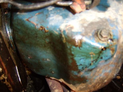 1989 Ford Bronco - Close-up of a grimy, oil-stained engine component painted in faded teal/blue, showing surface rust and a large hex bolt head on the right s…