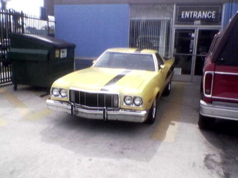 1988 Ford Bronco - A yellow coupe with racing stripes is parked in front of a building entrance next to a red pickup truck.