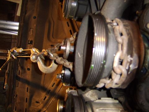 1989 Ford Bronco - A rotated close-up of an engine's front, showing a large ribbed serpentine crankshaft pulley/harmonic balancer with a rusty steel lift chai…