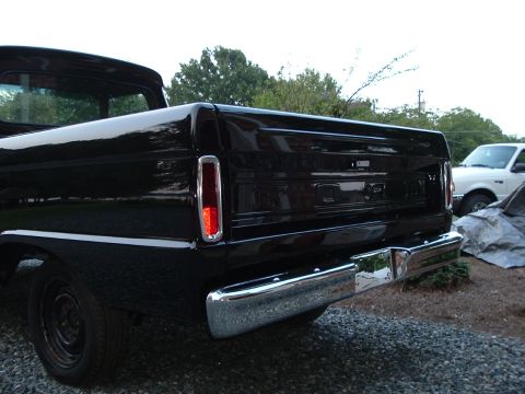 1966 Ford F-100 - The rear of a black pickup truck is shown with a chrome bumper and a white pickup truck in the background.