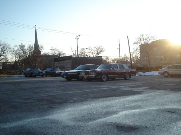 maroon sedan parked next to a black sedan, with other cars and a church steeple in the background