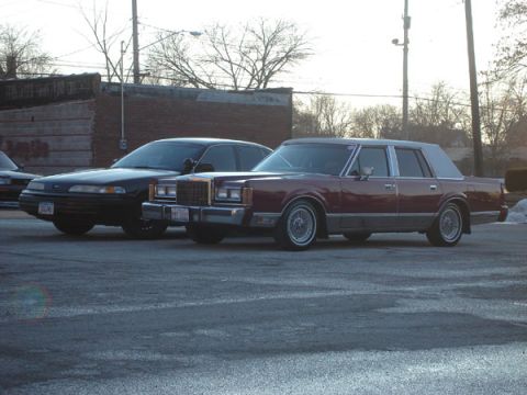 1992 Ford Crown Victoria - A maroon Lincoln Town Car and a black sedan are parked next to each other on a paved surface.