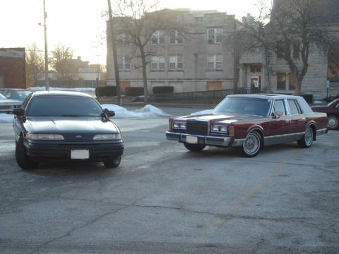 1992 Ford Crown Victoria - Two sedans are parked in a lot with buildings in the background.