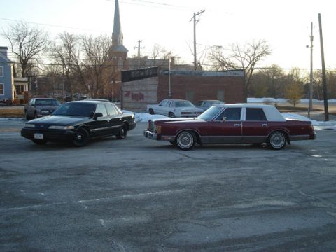 1992 Ford Crown Victoria - Two older model sedans are parked in a lot with a church steeple in the background.