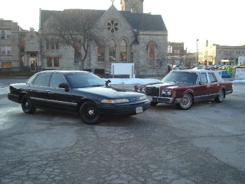 1992 Ford Crown Victoria - Two large sedans are parked in a lot with a church-like building in the background.