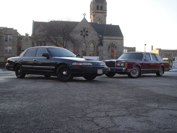 Black 1992 Ford Crown Victoria parked next to a maroon Lincoln Town Car, front 3/4 view