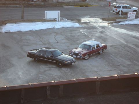1992 Ford Crown Victoria - A black 1992 Ford Crown Victoria is parked alongside a maroon sedan on a lot with patches of snow.