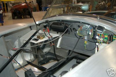 1965 Shelby Cobra - The exposed wiring and components in the engine bay of a car are visible, with another vehicle in the background.