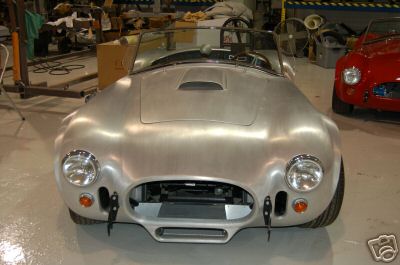 1965 Shelby Cobra - An unpainted aluminum 1965 Shelby Cobra is shown from the front, with another red Cobra visible in the background.