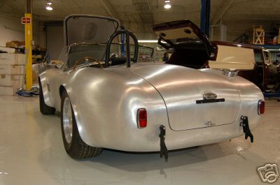 1965 Shelby Cobra - Rear view of an unfinished aluminum roadster with its doors and trunk open, parked in a workshop.